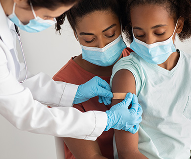 A masked medical assistant places a bandage on the arm of a child after vaccination.