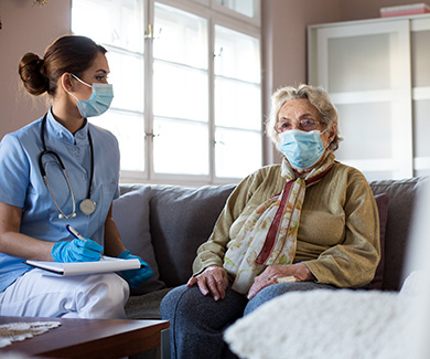 A masked medical assistant takes information from an elderly female patient.