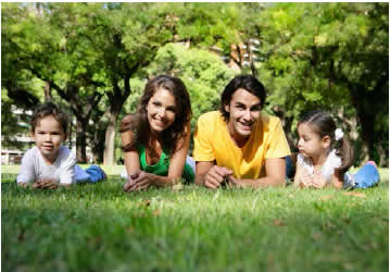 Photograph shows a happy family of two parents and two small children lying on the grass.